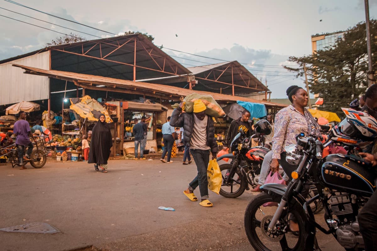 A busy open-air market street in Arusha, Tanzania, with vendors, shoppers and boda-boda motorcycles at dusk.
