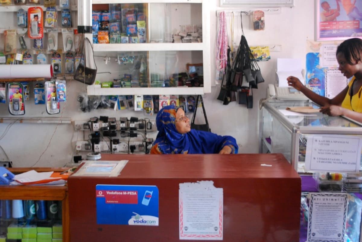 A Vodacom M-Pesa mobile-money agent inside a small shop in Tanzania, with the 'M-Pesa available here' sign on the counter.